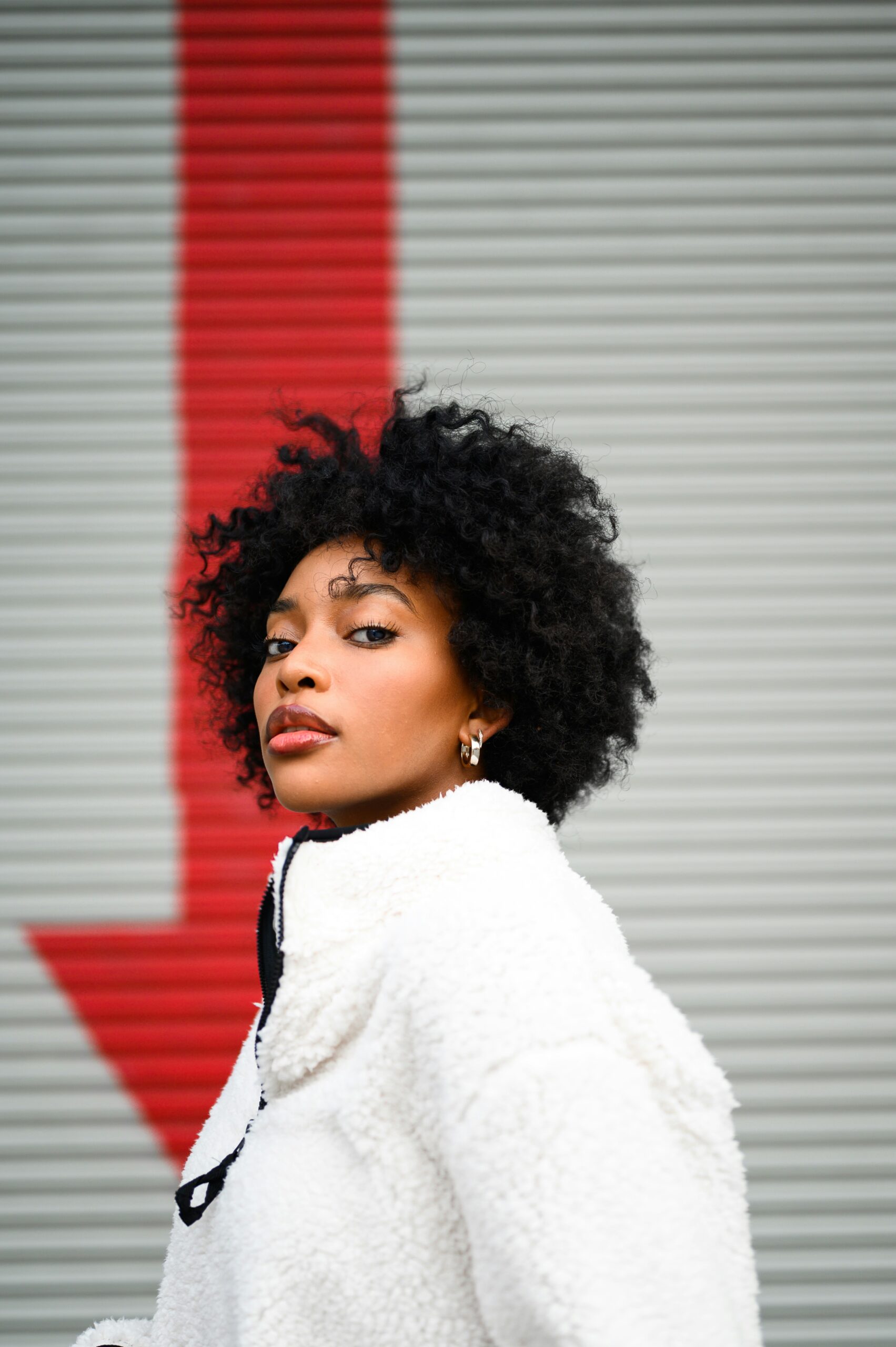 a fashion content creator with curly hair wearing jacket, posing in front of an urban street background with a red arrow design